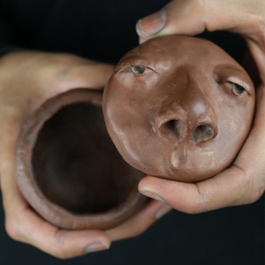Brown resin cast sculptural bowl featuring eyes and a nose, with the lid off to show the container behind it.