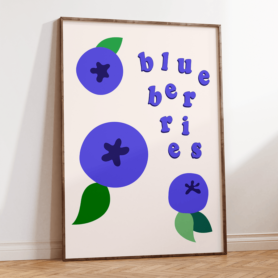 Frames art print of cartoon colour block purpley blue blueberries with green leaves and blue text saying ‘blueberries’ in floating letters. Frame is thin dark wood and is on a wooden floor, resting against a white wall.