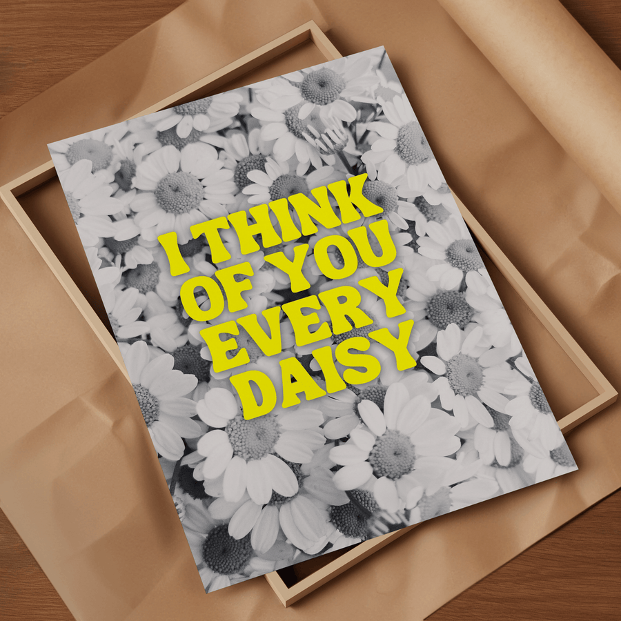 Black and white photograph of daisies, with neon yellow text overlay saying ‘I think of you every daisy’. Art print laid on top of an empty frame on top of brown packing paper.