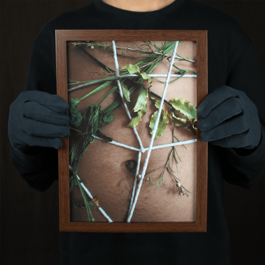 Photograph of a latino man’s belly and belly button with white string tied around his body with flowers intertwined, in a dark wooden frame.