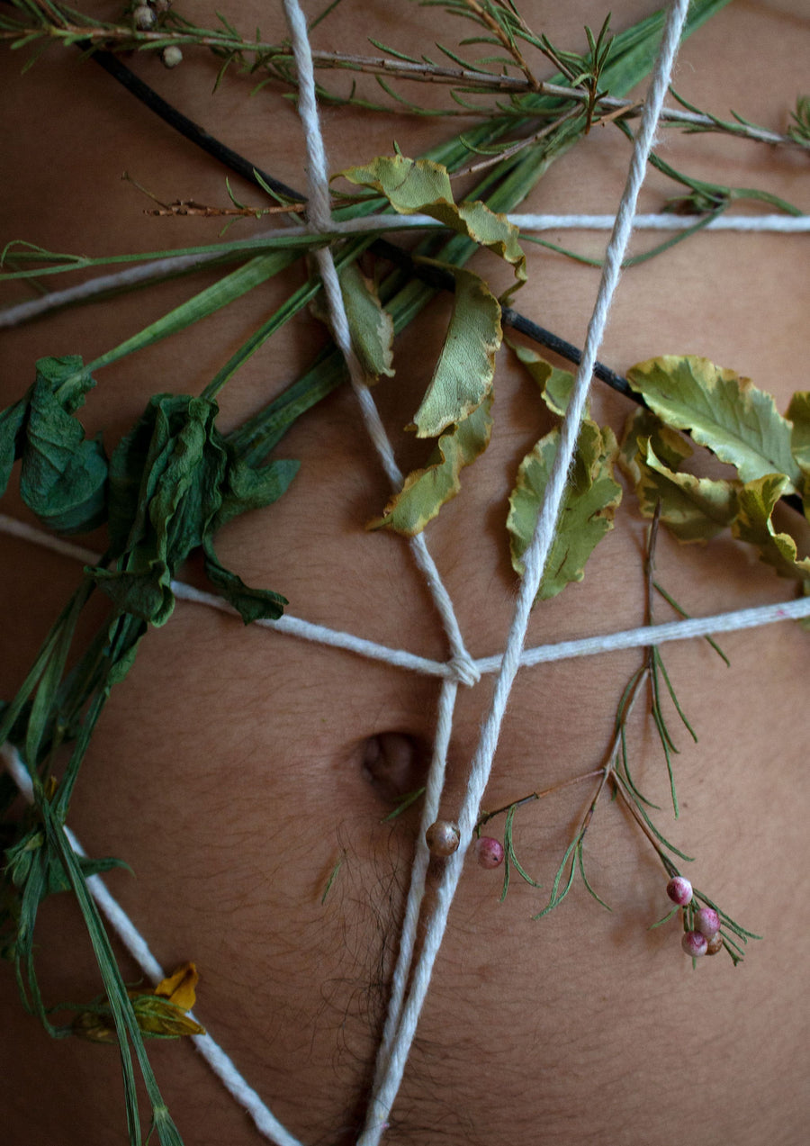 Photograph of a latino man’s belly and belly button with white string tied around his body with flowers intertwined, in a dark wooden frame.