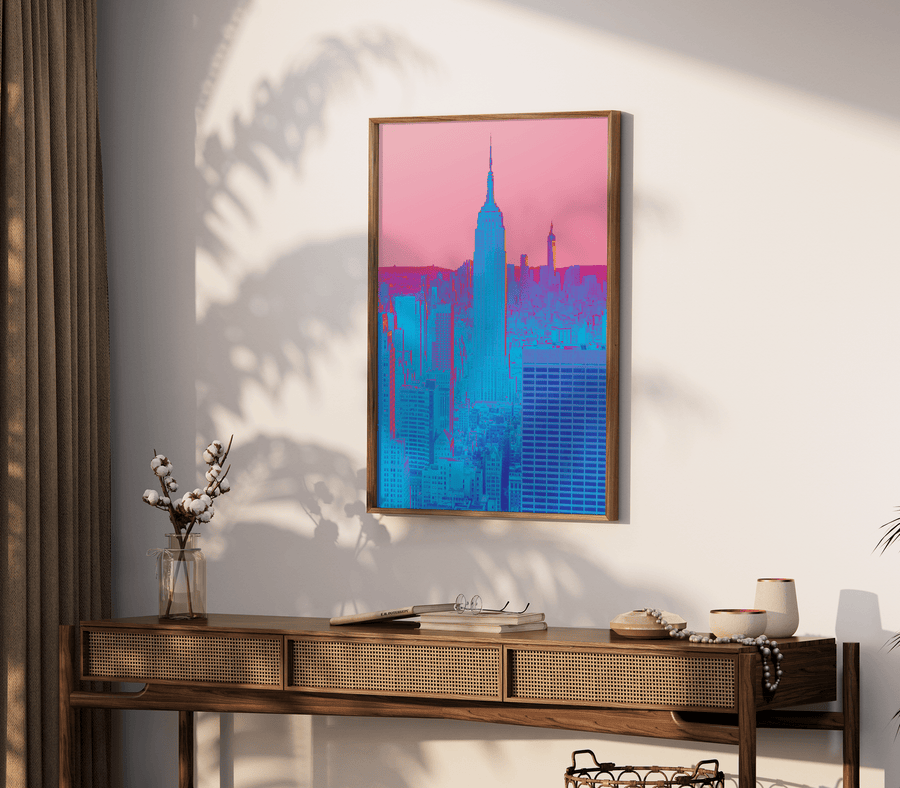 Art print of hyper colourful, experimental photography print of New York City in neon blue and pink, set against a faded pink sky. Print is in a thin wooden frame, on an off-white wall above a console table.