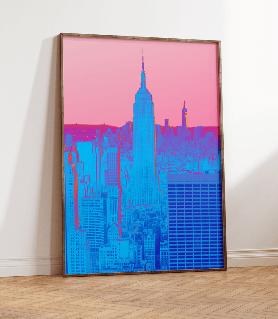 Hyper colourful, experimental photography print of New York City in neon blue and pink, set against a faded pink sky. Frame is thin dark wood and is on a wooden floor, resting against a white wall.