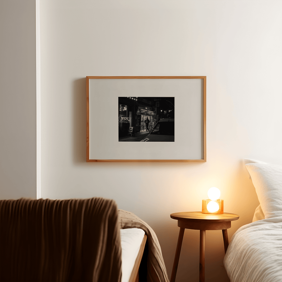 Black and white photograph by Yasmin Centeno, of a person stood outside a convenience store at night. Art print is in a light wood frame with a large off-white border on a white wall in a room with mid-century style furniture.