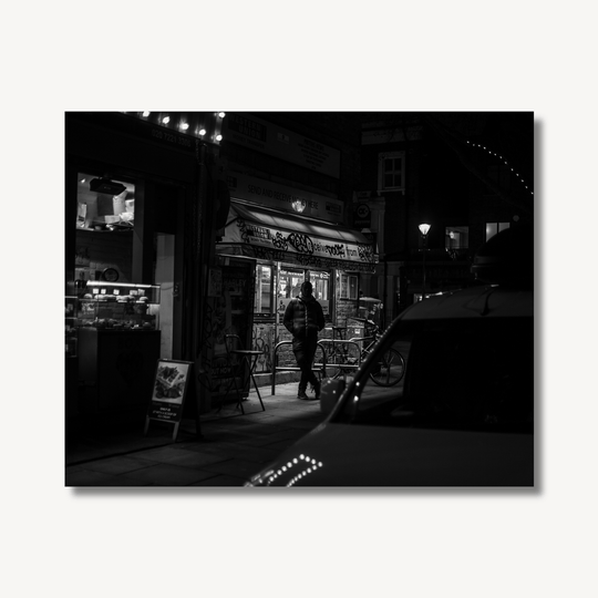 Black and white photograph of a person stood outside a convenience store at night.