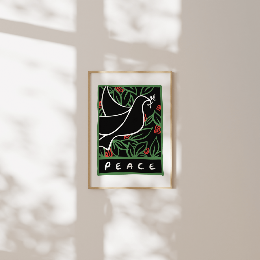 A white line drawing art print of a dove holding an olive branch, surrounded by green leaves and red flowers with the word 'Peace' at the bottom, displayed in a thin pale wooden frame. The frame is hung on a white wall, with soft window light and shadows casting across its surface.