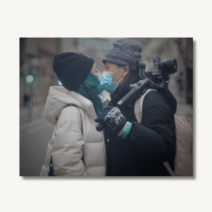 Two queer Chinese people kissing in the street. Both wearing puffy coats and beanie hats, while wearing blue surgical face masks. One is carrying a camera.