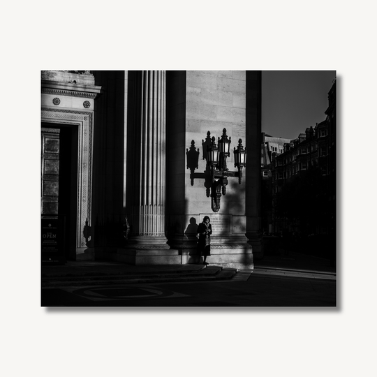 Black and white photograph of a woman stood waiting by a large building with large shadows cast across the scene by the sunset shining through the cityscape.