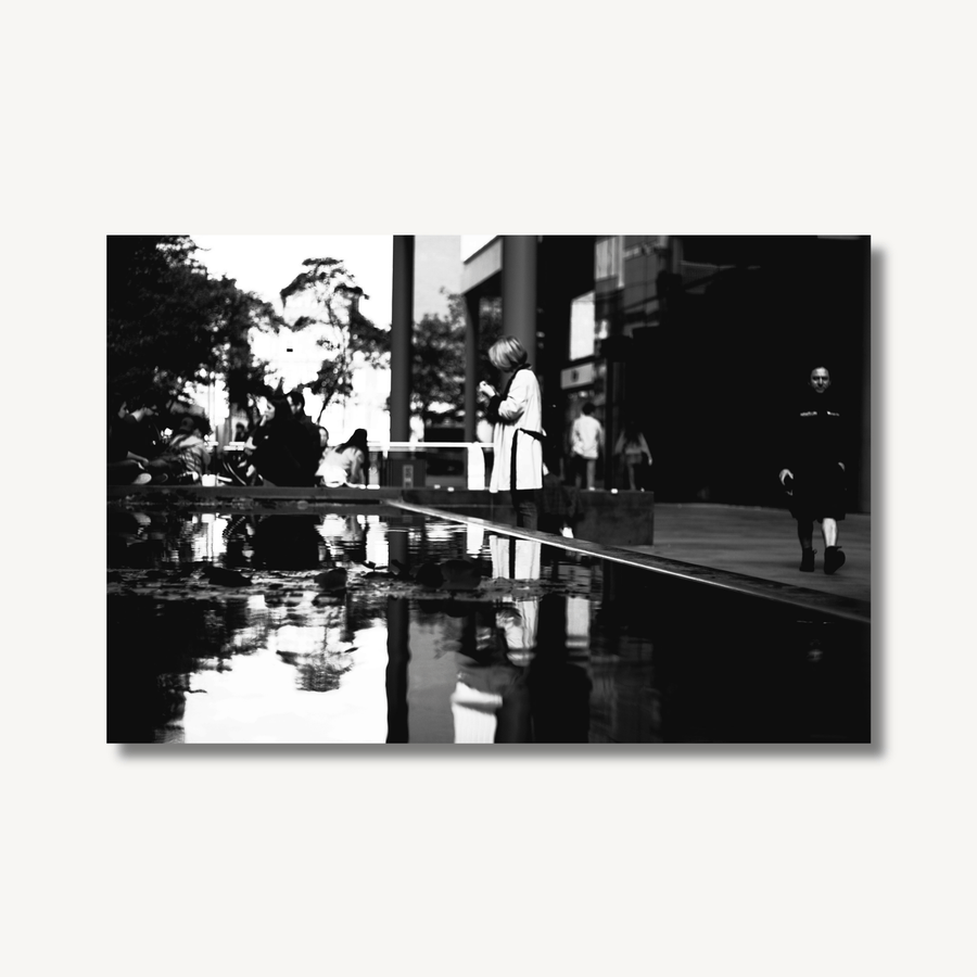 High contrast black and white photograph of a woman stood in front of a man-made pond in between London office buildings, showing a clear reflection in the still water of the pond.