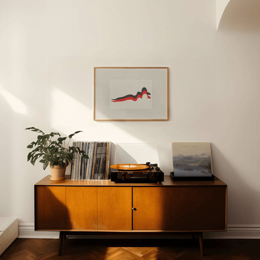 Paper collage of a woman’s figure lying down, using cut out black and red paper on a white background. In a large wooden frame on a white wall above a mid-century style side board with a record player on top.