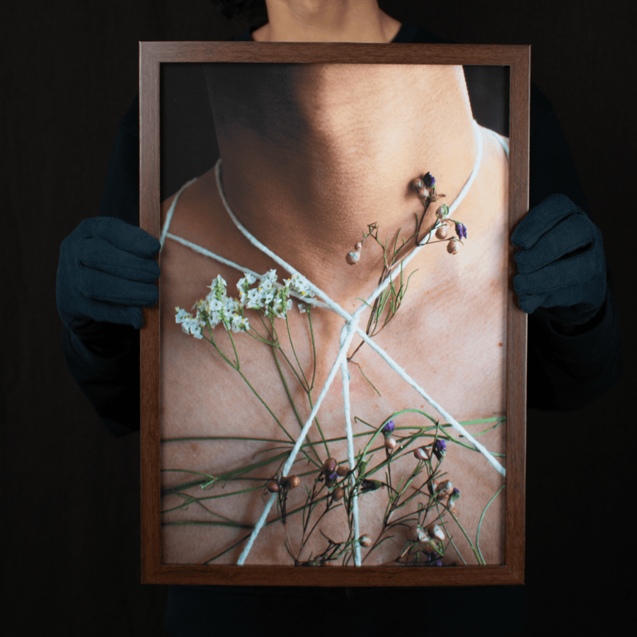 Photograph of a latino man’s neck with white string tied around his body with flowers intertwined, in a dark wooden frame.