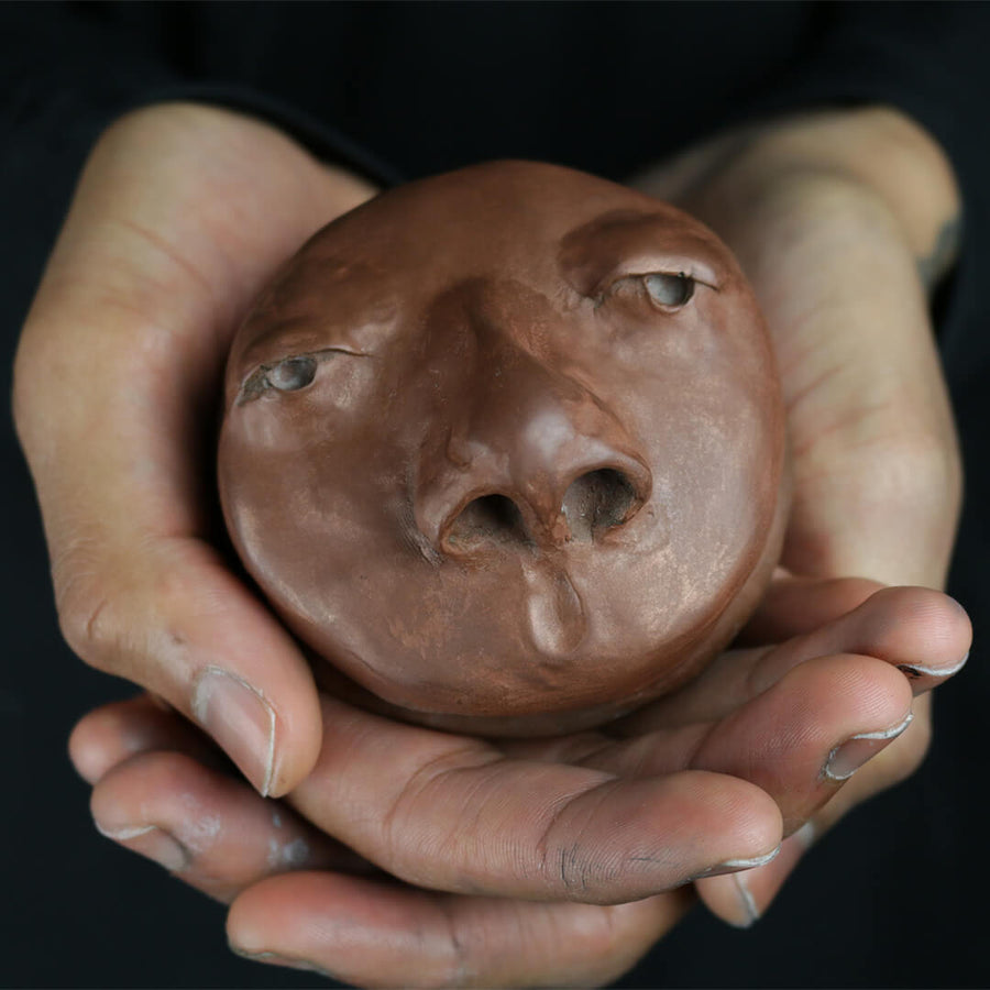 Brown resin cast sculptural bowl featuring eyes and a nose, being held in a person’s hands to show scale.