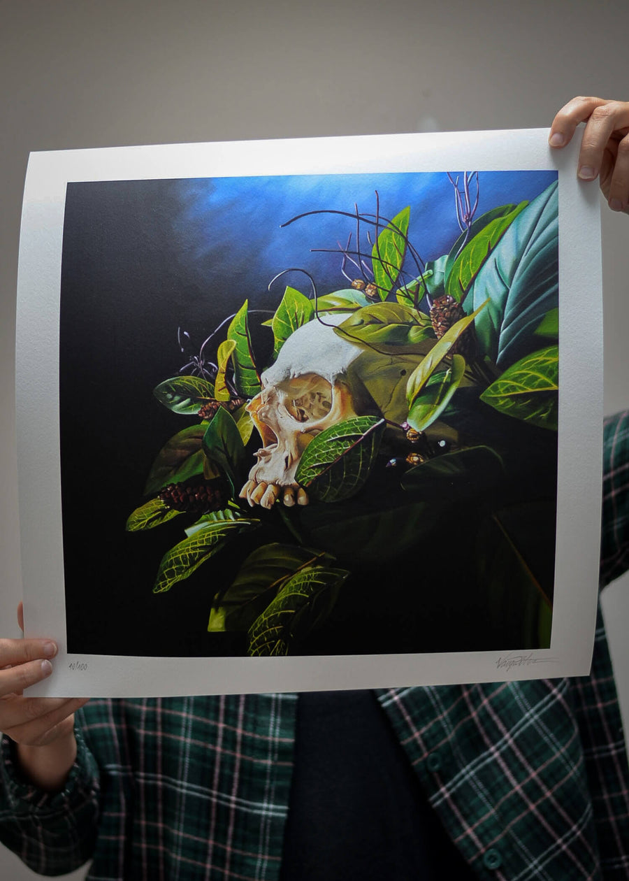 Tattoo artist, Tibor Varga, holding a print of his artwork ‘Death of Paradise’ – a traditional style oil painting of a skull with leaves and flowers, on a blue background.