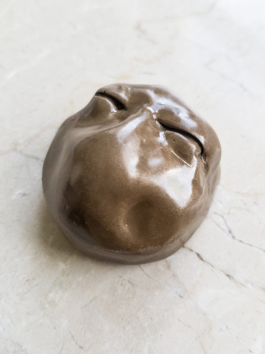 Brass coloured resin cast sculpture of a face with no mouth or nose with a focus on the eyes which are closed tight.