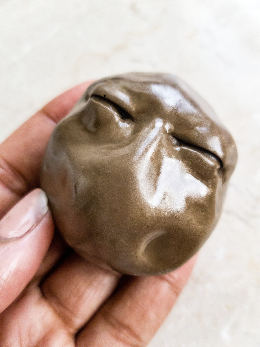Close up of black woman’s hand holding a small brass coloured resin cast sculpture of a face with no mouth or nose with a focus on the eyes which are closed tight.