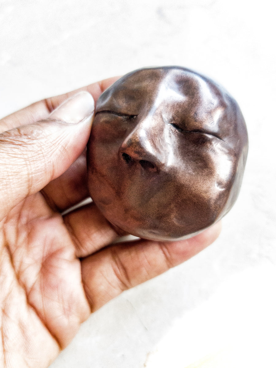 Close up of black woman’s hand holding a small Copper/bronze coloured resin cast sculpture of a face with no mouth, with closed eyes.