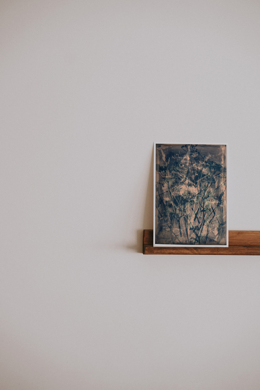 Plain wall with wooden shelf off to the side of the image with a framed artwork on. Artwork shows a dark blue/black and pale amber cyanotype print of flowers.