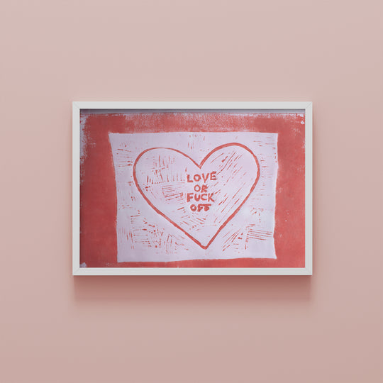 Lino print in dark pink on pale pink paper of a rough square with a heart in the centre with the words ‘love or fuck off’ in the middle. In a think white frame on a pale pink background.