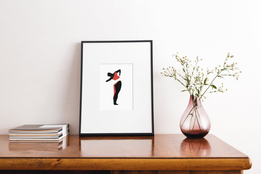 Paper collage of a woman’s figure, using cut out black and red paper on a white background. In a thin black frame on a wooden table next to a stack of magazines and a vase with wild flowers.