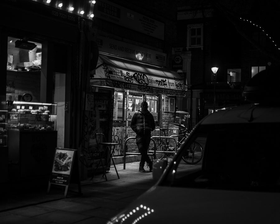 Black and white photograph of a person stood outside a convenience store at night.